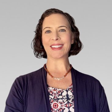 Smiling woman with short curly hair wearing a navy cardigan and patterned blouse against a gray gradient backdrop, necklace visible.