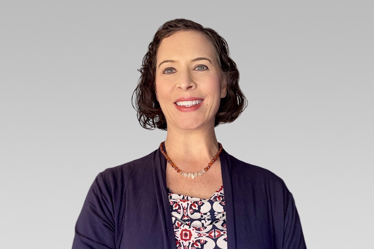 Kerri Fair headshot Large Smiling woman with short curly hair wearing a navy cardigan and patterned blouse against a gray gradient backdrop, necklace visible.